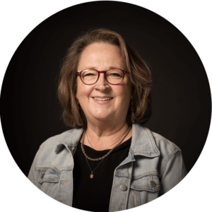 Smiling middle-aged woman with shoulder-length brown hair wearing red glasses, a light denim jacket over a black top, and layered necklaces, posing against a dark background.