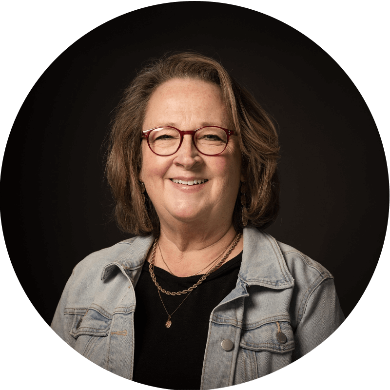 Smiling middle-aged woman with shoulder-length brown hair wearing red glasses, a light denim jacket over a black top, and layered necklaces, posing against a dark background.