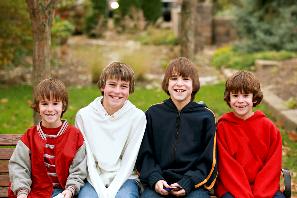 Four young boys sitting on a wooden bench outdoors, smiling warmly with a natural, green backdrop of trees and bushes.