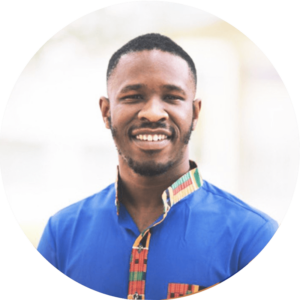 Smiling man wearing a vibrant blue shirt with colorful patterned trim on the collar and chest, standing against a light, blurred background.