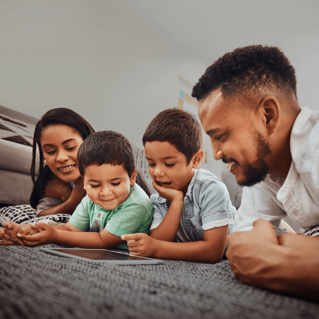 A family of four, including a mother, father, and two young boys, lies on the floor together, smiling and interacting with a tablet in a warm, relaxed home setting.