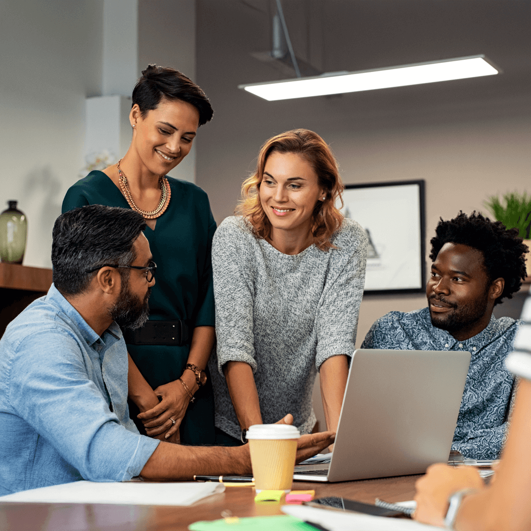 A diverse group of professionals collaborating in an office, gathered around a laptop with smiling and engaged expressions.