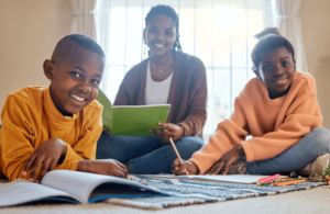 A mother sits on the floor with her two children, smiling and engaging in a creative activity with colored pencils and notebooks.