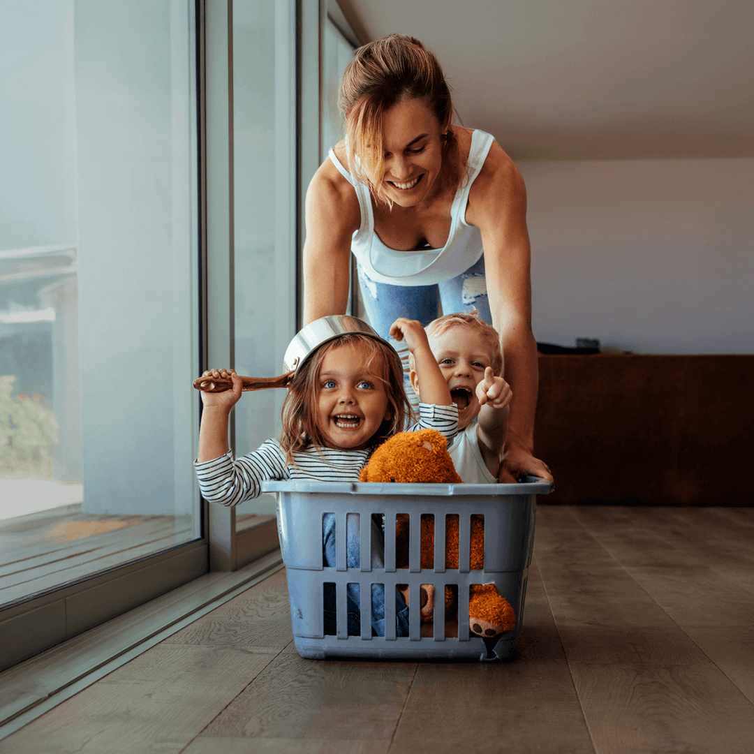 A playful moment of a smiling mother pushing two laughing children in a laundry basket, one child wearing a saucepan as a helmet and holding a wooden spoon, with a bright home setting in the background.