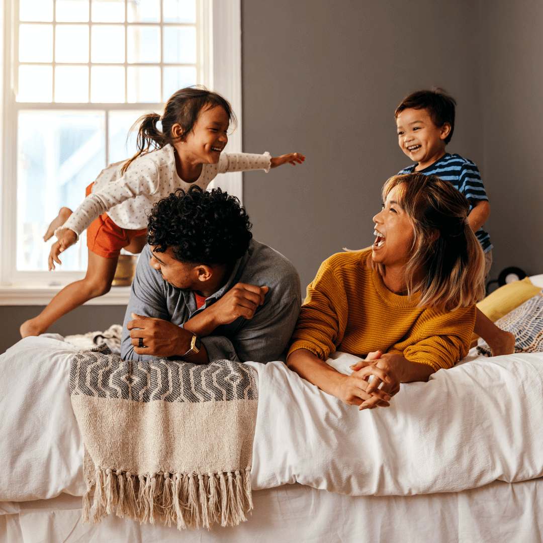 A joyful family of four playing on a bed, with parents laughing and children energetically jumping and interacting.