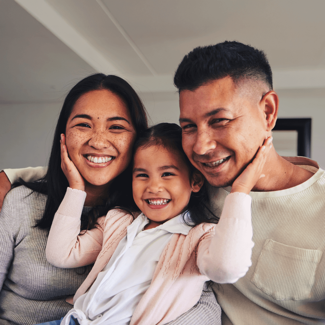 A joyful family of three, including a mother, father, and young daughter, sitting close together and smiling warmly while embracing each other in a bright, cozy home.