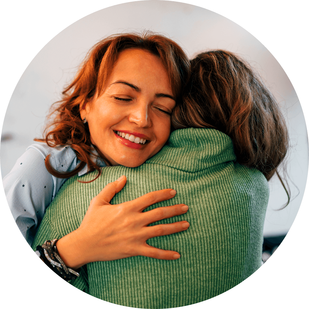 A smiling mother with red hair warmly hugs her teen girl wearing a green sweater, radiating comfort and connection.