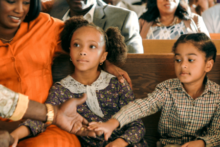 Two young children sitting in a church pew, one reaching out to shake an adult's hand while the other looks on. They are surrounded by warmly dressed adults in a welcoming environment.