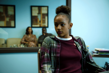 A young girl sits in an office chair, looking to the side with a guarded expression. In the background, two adults sit in a waiting area, slightly blurred, creating a sense of distance.