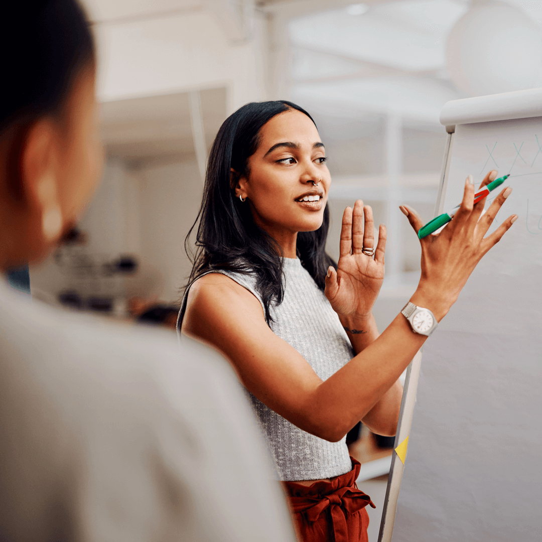 A professional woman presenting at a whiteboard, holding a marker and gesturing confidently while speaking to an engaged audience.