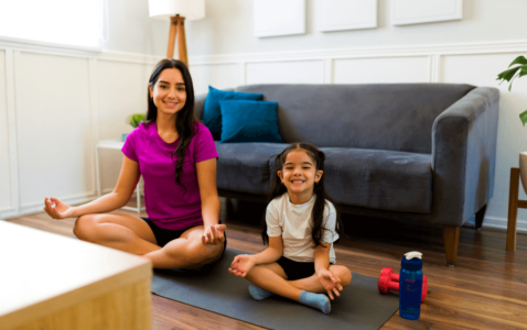 A mother and her young daughter sitting cross-legged on yoga mats, smiling and practicing mindfulness in a cozy living room with a gray couch, blue pillows, and wooden floors.