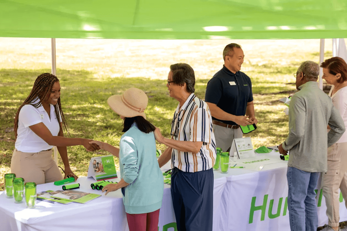 A community engagement event under a green tent, where Humana Foundation representatives interact with attendees at an outdoor booth. A smiling woman in a white polo shirt shakes hands with a visitor while another representative in a navy polo assists others. The table is covered with promotional materials, including green water bottles and brochures.