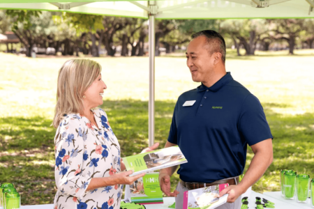 A Humana Foundation representative in a navy polo shirt shares informational brochures with a smiling woman at an outdoor event booth, surrounded by branded materials and green promotional items.