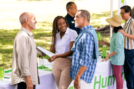 A Humana Foundation representative in a white polo shirt smiles while engaging in conversation with two older men at an outdoor event. The booth is covered with promotional materials, including brochures, water bottles, and branded merchandise. Other attendees interact in the background under a green tent.