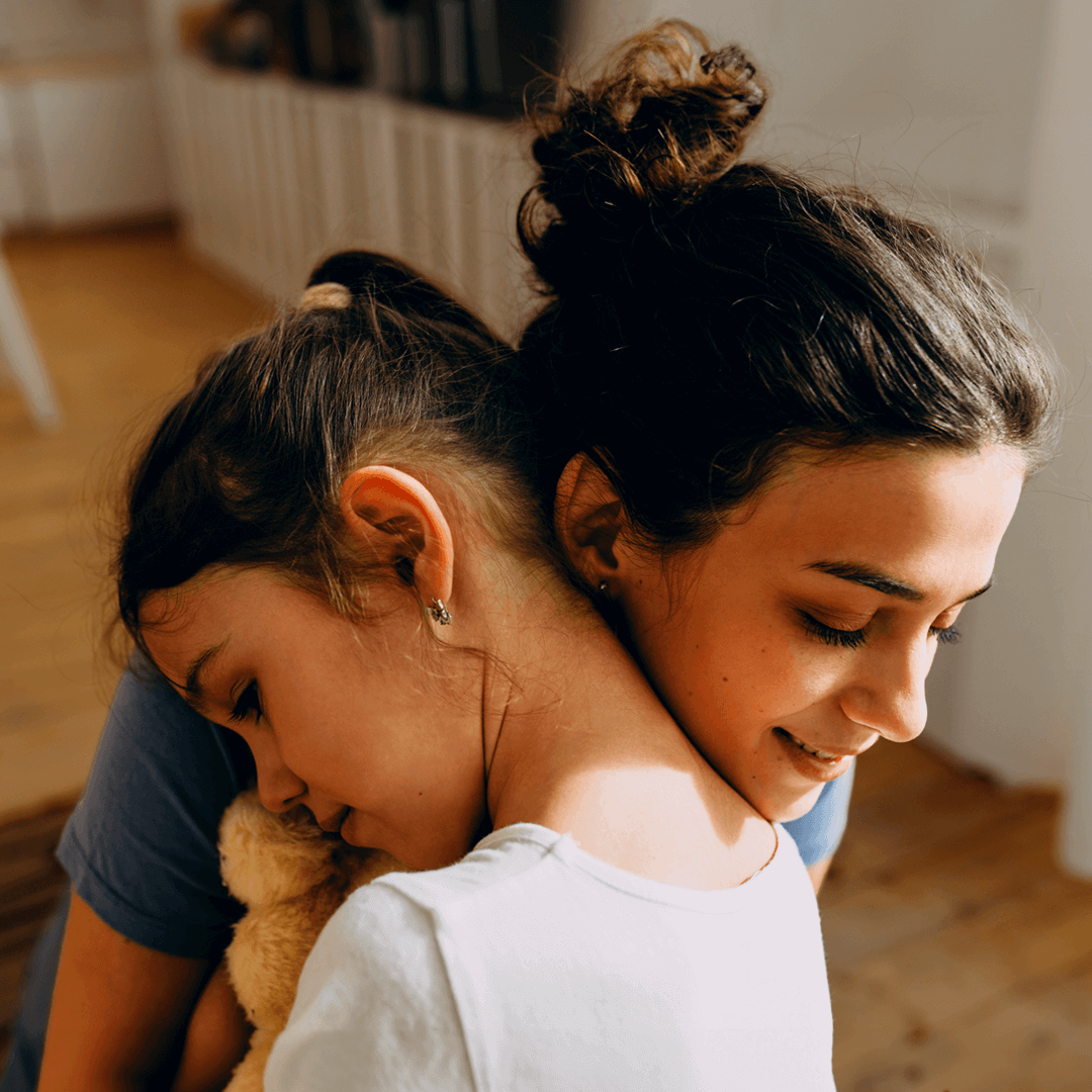 A young girl embraces an older girl with a soft smile, holding a teddy bear close. The warm lighting highlights their connection and sense of comfort.