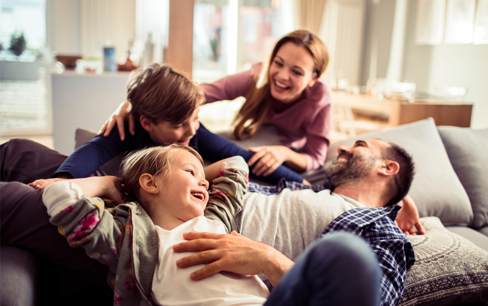 A joyful family moment, with parents and two children laughing together on a cozy couch in a bright living room.