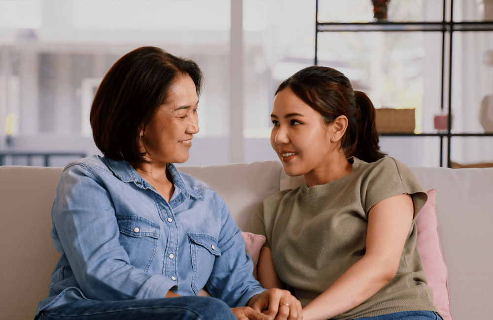 A mother and daughter sit closely on a couch, holding hands and smiling warmly at each other, conveying a sense of connection and understanding.