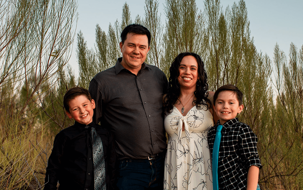 A family of four standing outdoors with tall green plants in the background. The mother, wearing a white floral dress, and the father, in a dark patterned shirt, stand in the center with their two young sons on either side, both wearing ties and smiling warmly.