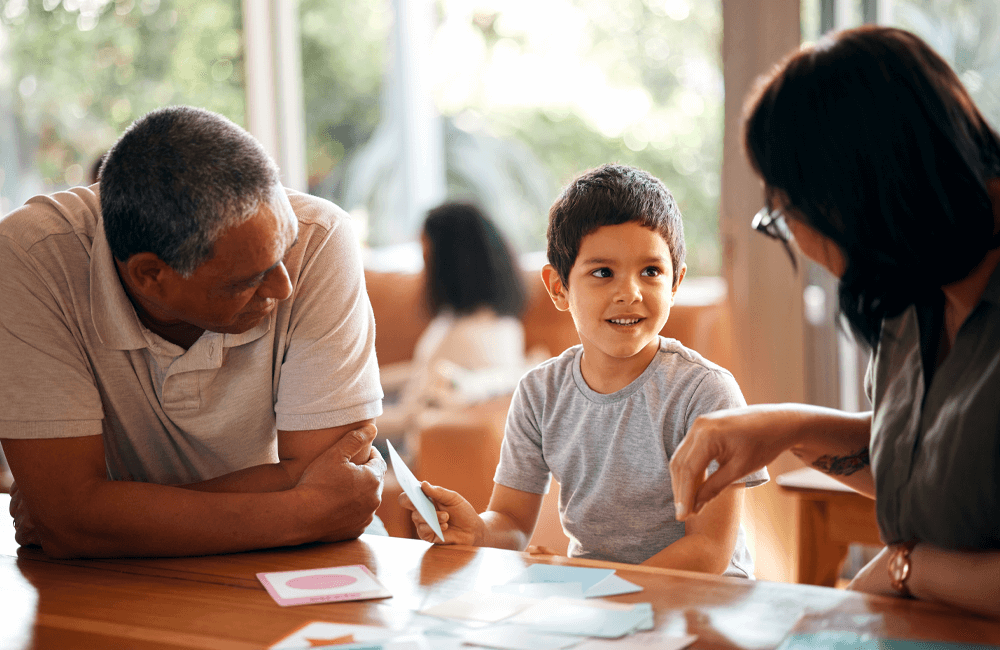 A young boy sitting at a table with two adults, smiling and engaging in a positive, caring conversation. Bright natural light and a warm atmosphere suggest a safe, nurturing environment.