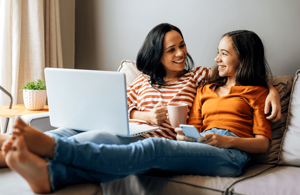 Mother and teenage daughter smiling and relaxing on a couch with a laptop and smartphone, sharing a warm moment together.