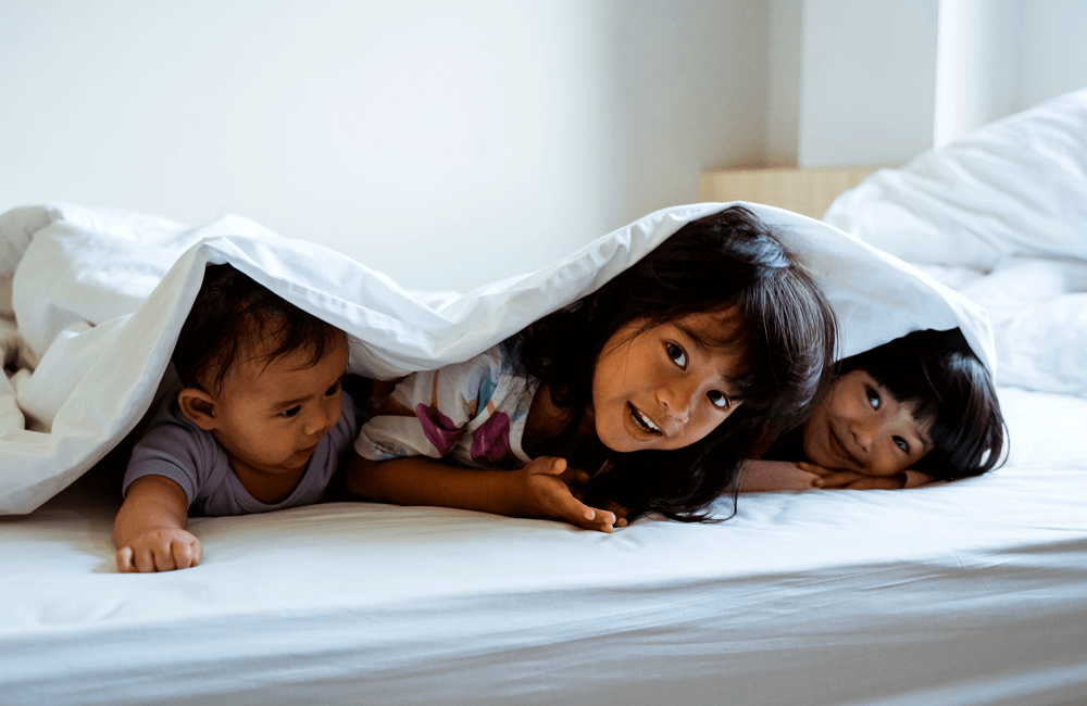 Three young children lying under a white blanket on a bed, smiling and playing together.